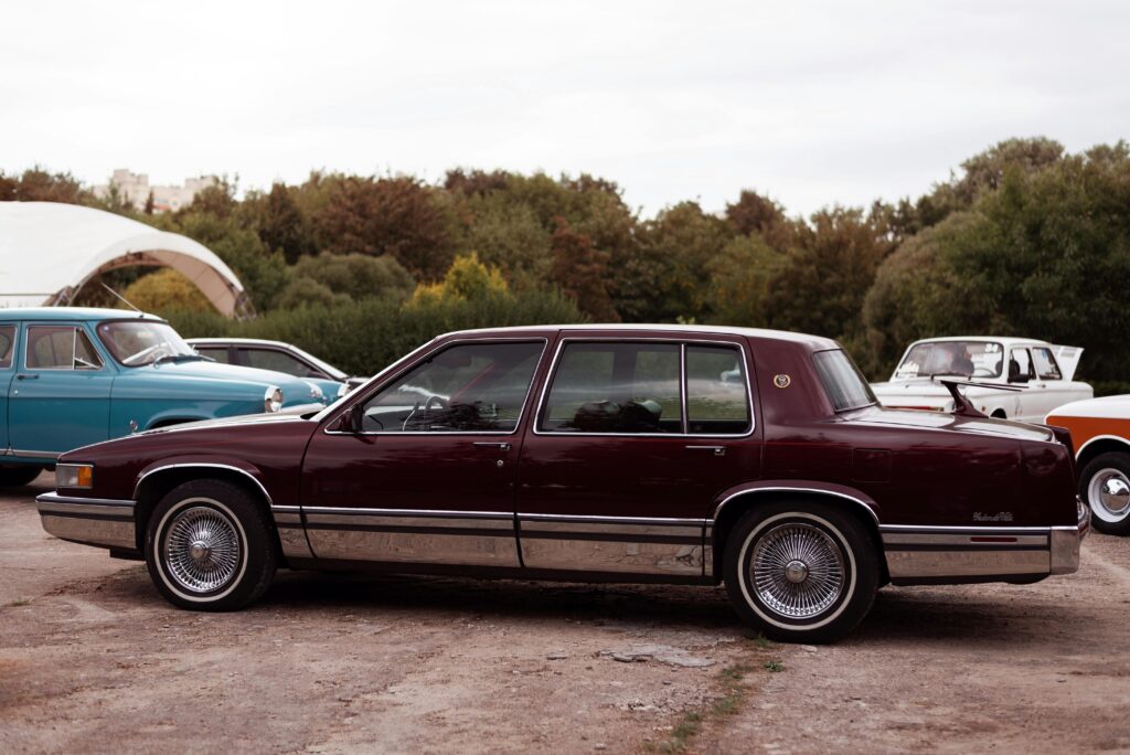 Classic maroon Cadillac De Ville parked against a lush green backdrop featuring other vintage cars.